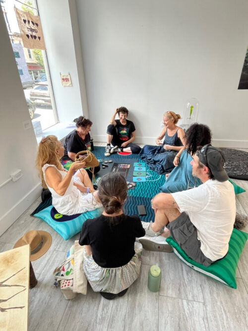 Six people including Abigail Simon and Marina Zurkow sit in a circle on the floor of a gallery playing The Iceberg